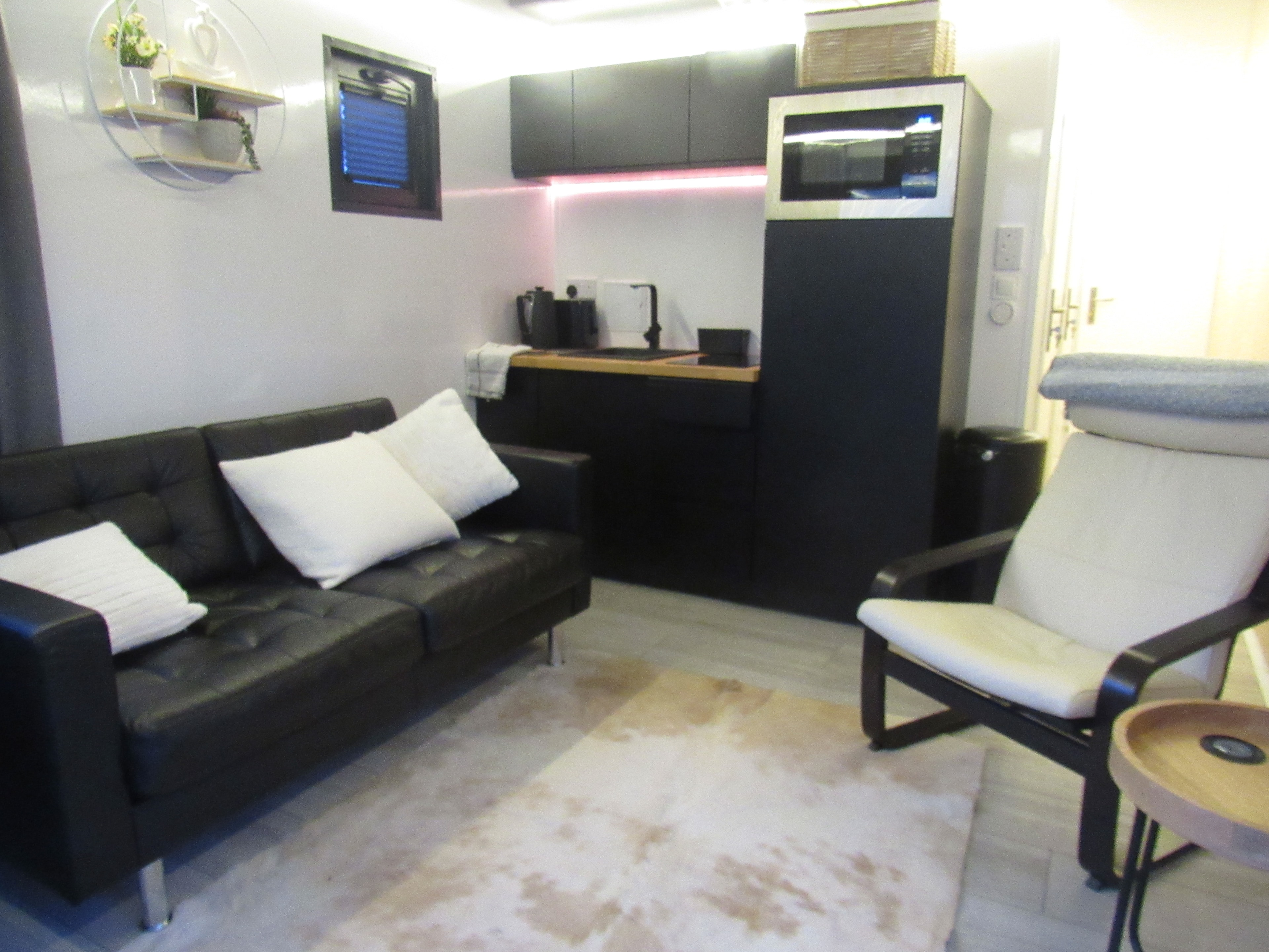 Living room overview showing black leather sofa, armchair, kitchenette and pink LED lighting